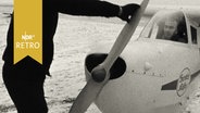Mann startet Propeller eines Kleinflugzeugs per Hand auf einem schneebedeckten Feld (1962)  