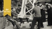 Bauarbeiter bringen an Kran hängenden Hochspannungsmast in Position (1962)  