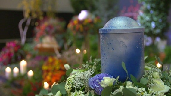 Im Vordergrund eine blaue Urne mit einem Blumengesteck drumherum. Unscharf im Hintergrund weitere Blumenarrangements und Särge. Aufnahme auf einer Messe. © NDR 