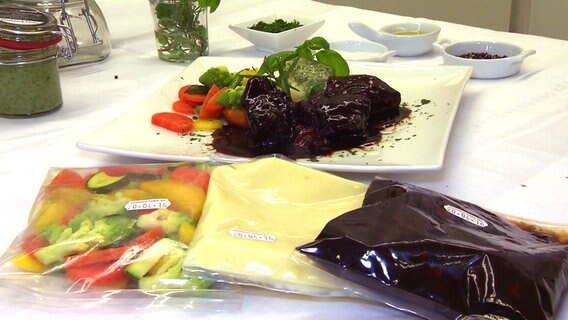 Beispiel für High Convenience Food: Auf einem Teller angerichtet sind Kalbsbäckchen, gemischtes Gemüse und Kartoffelstampf. Davor liegen durchsichtige Plastikbeutel mit den Bestandteilen des Gerichts. © NDR 