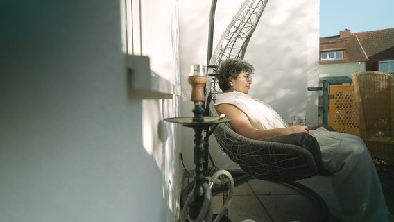Eine Protagonistin aus der Doku "Sieben Jahre Deutschland" sitzt in einem Hängestuhl auf ihrem Balkon in der Sonne. © NDR Foto: Alexandra Bidian