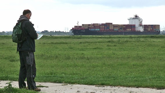Vogelkundler Markus Risch steht auf einem Feldweg der Haseldorfer Marsch. Im Hintergrund zieht ein Containerschiff vorbei. © NDR 