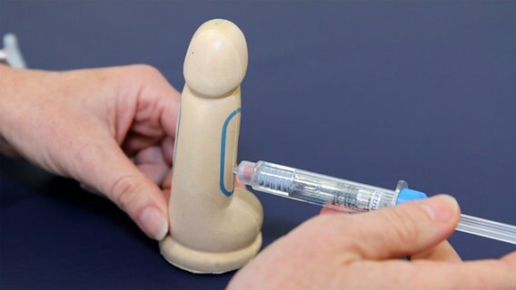 Mithilfe eines Penis-Dummys wird gezeigt, wie ein Medikament ins Glied gespritzt wird. © NDR 
