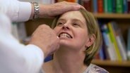 Eine Kopfschmerzpatientin hat den Kopf in den Nacken gelegt und zeigt ihre Zähne, damit der vor ihr stehende Arzt den Stand des Kiefers beurteilen kann. © NDR 