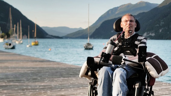Harald Mayer sitzt in seinem Rollstuhl. Dieser steht auf einem Steg an einem See mit Bergen im Hintergrund. © NDR 