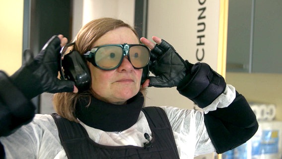 Frau steckt in einem Alterssimulationsanzug und Gewichte und Manschetten an den Armen, Kopfhörer sowie eine die Sicht einschränkende Brille. © NDR 