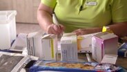Ein Tisch mit mehreren offenen Medikamentenpackungen. Dahinter sitzt eine Frau, von der nur der Oberkörper zu sehen ist, und macht Notizen. © NDR 