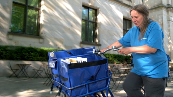 Eine Briefträgerin von einem anderen Dienst als der Post (blaue Uniform statt gelb) schiebt ihr Fahrrad mit der Brieftasche und schaut nach unten. © NDR 