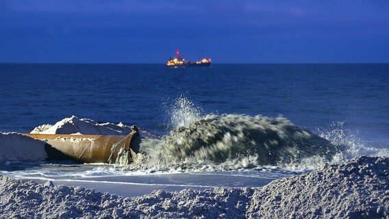 Sandvorspülung am Strand von Sylt mit Blick aufs Meer. Parallel zur Wasserlinie liegt ein großes Rohr aus dem ein Sand-Wasser-Gemisch sprudelt. Im Hintergrund, ist das beleuchtete Baggerschiff zu erkennen. © NDR 