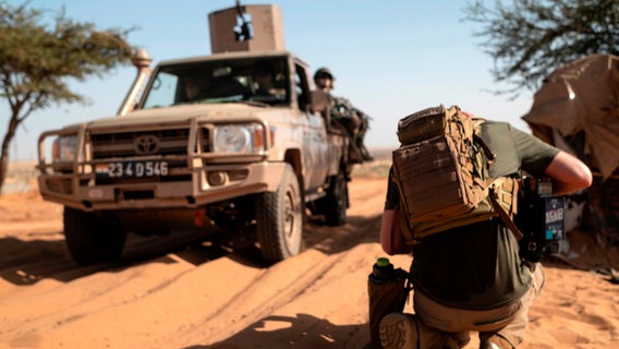 Aufnahme einer Drehsituation in der Wüste: Pickup auf Sandspur im Hintergrund. Davor kniet der Kameramann, der von hinten zu sehen ist, und filmt den Wagen, auf dessen Ladefläche ein Soldat zu sehen ist. © NDR/Martin Ross 