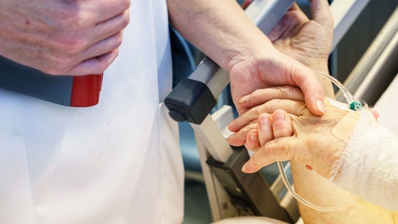 Pflegerin mit Patientendokumentation im Arm hält mit der anderen Hand, die Hand eines alten Patienten mit einem Schlauch im Handrücken. © IMAGO/epd 