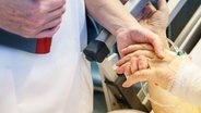 Pflegerin mit Patientendokumentation im Arm hält mit der anderen Hand, die Hand eines alten Patienten mit einem Schlauch im Handrücken. © IMAGO/epd 