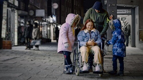 Nächtliche Straßenszene: Eine Frau mit Prothesen an den Beinen sind im Rollstuhl und schaut auf ihr Handy. Links und rechts stehen ihre Kinder, hinter ihr ihr Mann. © NDR Foto: Lennart Banholzer / Alexandra Bidian