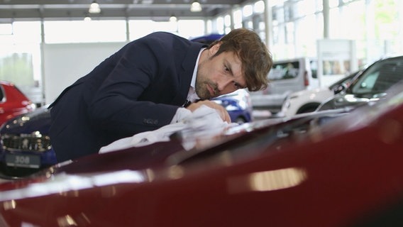 Ein Mann im Anzug poliert ein rotes Auto. © NDR/7 Tage 