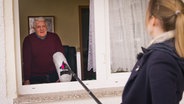Eine junge Frau hält ein langes Mikrofon in ein Fenster. Ein älterer Mann spricht hinein. © NDR/7 Tage 