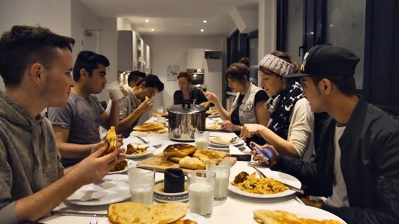 Zahlreiche Menschen sitzen an einem Tisch und essen gemeinsam. © NDR 