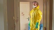 Ein Mann in gelbem Schutzanzug und Atemmaske steht in einem Raum voller Blutspritzer an den Wänden. © NDR 