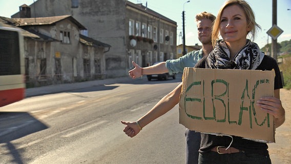 Eine Frau und ein Mann stehen mit einem Pappschild an der Straße und halten die Daumen hoch. © NDR 