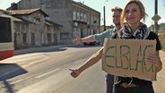 Eine Frau und ein Mann stehen mit einem Pappschild an der Straße und halten die Daumen hoch. © NDR 