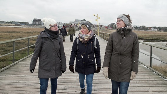 Drei junge Frauen gehen in Winterjacken und mit Mützen einen Steg am Strand entlang. © NDR/7Tage 