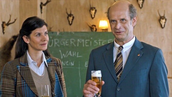 Szenenbild aus der sechsteiligen Komödie "Da is' ja nix": Eine Frau und ein Mann (Gerda & Frank Petersen) stehen mit einem Getränk in der Hand vor einer Tafel. © NDR 