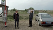 Szenenbild aus der sechsteiligen Komödie "Da is' ja nix": Zwei Männner stehen an einer Bushaltestelle, im Hintergrund ein See. © NDR 
