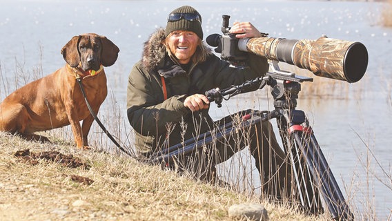 Andreas Kieling posiert mit einem Hund und mit einer Kamera auf einem Stativ in der Natur. © NATIONAL GEOGRAPHIC/Andreas Kieling 