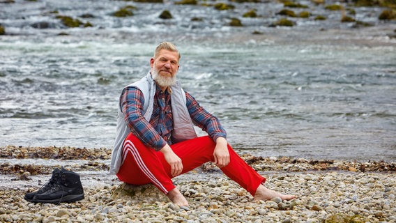 Ein Mann sitzt vor einem Fluss auf dem Kiesbett. © ZS - ein Verlag der Edel Verlagsgruppe 