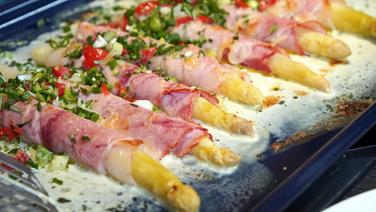 Asparagus and ham rolls with cheese, diced tomatoes and herbs on a baking sheet.  © NDR Photo: Florian Kruck