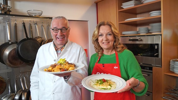 Rainer Sass und Bettina Tietjen stehen in einer Küche und halten Teller mit Nudelgerichten in die Kamera. © NDR/dmfilm/Florian Kruck 