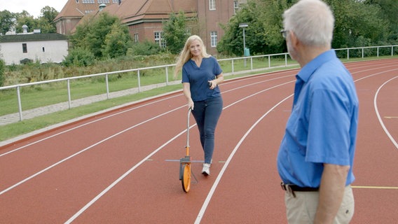 Melanie Hümmelgen läuft mit einem Entfernungsmesser auf dem Sportplatz, Patient Klaus G. schaut zu. © NDR/nonfictionplanet 