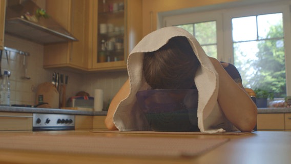 Frau sitzt am Tisch und hält den Kopf über eine Schüssel gebeugt, hat ein Handtuch übergelegt. © NDR 