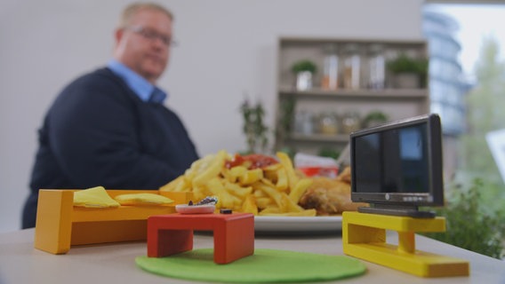 Ein etwass fülliger Mann blickt auf ein Arrangement aus Couch, Fernseher und Fast Food, das vor ihm im Fokus steht. © NDR Foto: Oliver Zydek/Moritz Schwarz