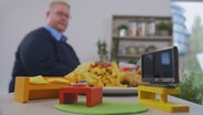 Ein etwass fülliger Mann blickt auf ein Arrangement aus Couch, Fernseher und Fast Food, das vor ihm im Fokus steht. © NDR Foto: Oliver Zydek/Moritz Schwarz