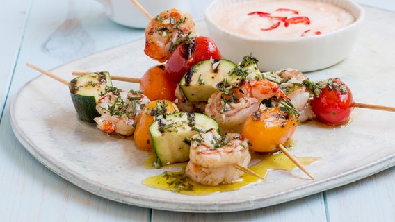 Three shrimp skewers are on a plate, next to a bowl of sauce.  © NDR Photo: Claudia Timmann