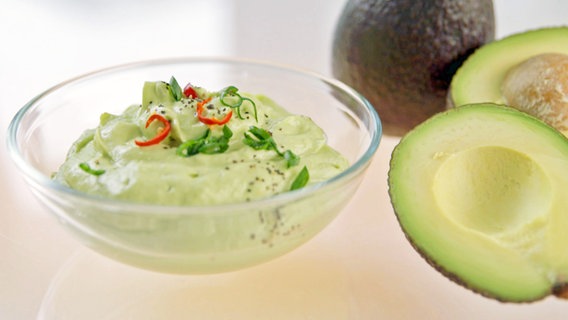 Ein Glasschälchen voll Guacamole steht auf einem Tisch, daneben einige Avocados. © NDR 