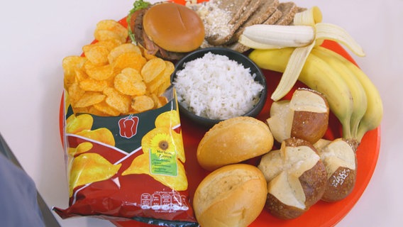 Brötchen, Laugengebäck, Chips, ein Whopper, Bananen, Brot und weißer Reis liegen angerichtet auf einem Teller. © NDR 