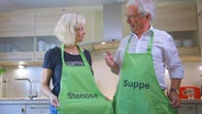 E-Docs Jörn Klasen spricht mit einer Patientin in deren Küche. Beide tragen Schürzen mit einer Aufschrift. Bei ihm steht "Suppe", bei ihr "Stenose". © NDR 