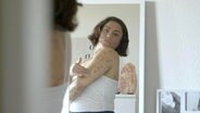 Eine Frau mit Neurodermitis am ganzen Körper cremt sich vor dem Spiegel ein. © NDR 
