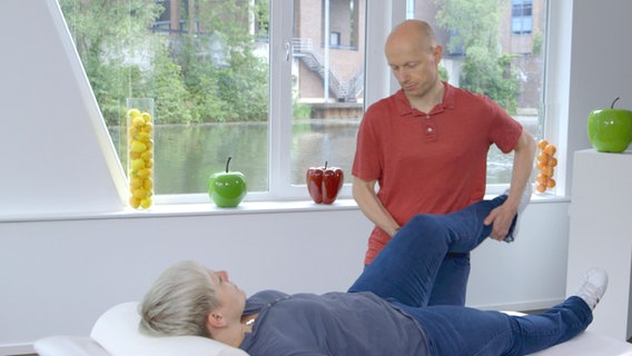 Bewegungs-Doc Helge Riepenhof hebt das gewinkelte Bein einer liegenden Patientin an. © NDR 