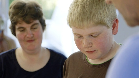 Übergewichtige Frau (links) und ihr Sohn stehen mit einem Arzt am Tisch. © NDR 