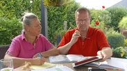 Paar sitzt am Gartentisch mit Stift, Ordner und Kochbüchern. © NDR/nonfictionplanet 