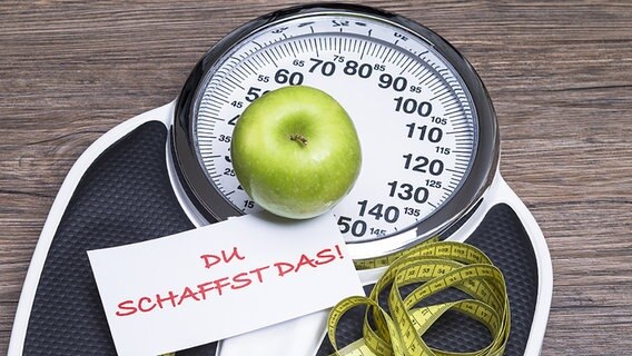 Personenwaage mit Apfel, Maßband und einem Zettel mit der Aufschrift: "Du schaffst das". © fotolia Foto: Ralf Kalytta