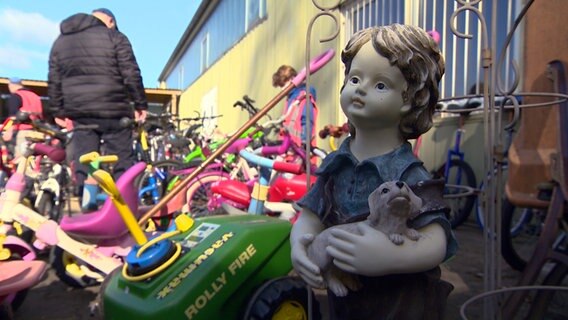 Eine Puppe steht auf einem Recyclinghof neben Kinderfahrrädern und anderen weggeworfenen Gegenständen. © NDR 