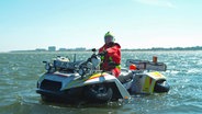 Ein Mann in einer Rettungsuniform und mit Helm sitzt auf einem Amphibienfahrzeug im Meer. © NDR/nonfictionplanet/Julian Ringer, honorarfrei Foto: Julian Ringer