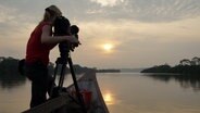 Eine Frau steht auf einem Floß und filmt den Sonnenuntergang über dem Fluss. © NDR/Doclights GmbH 