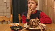 Ein Junge mit einer Zigarette im Mund und einem Hähnchen auf dem Teller.  