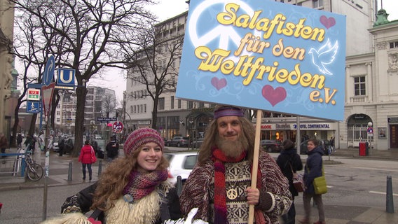 Tobias Schlegl mit langem Bart und Hippie-Klamotten hält ein Schild hoch mit der Aufschrift: "Salafisten für den Weltfrieden".  
