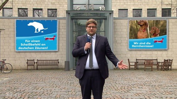 Reporter Henning Nasse steht vor zwei Plakaten der Partei Aufmerksamkeit für Dackel: "Für einen Scheißbefehl an deutschen Zäunen" und "Wir sind die Herrchenrasse".  Foto: Screenshot