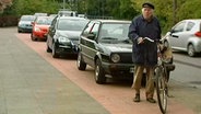 Mit Autos vollgeparkter Radweg, davor steht ein älterer Herr neben seinem Fahrad  